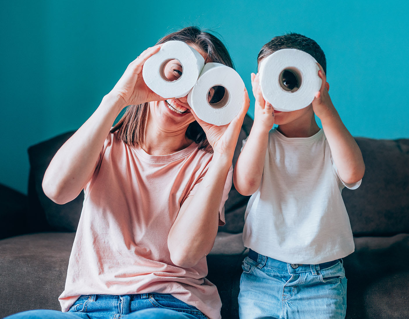 A woman and a child hold toilet paper rolls in front of their eyes, pretending to use them as binoculars, while sitting on a brown couch.